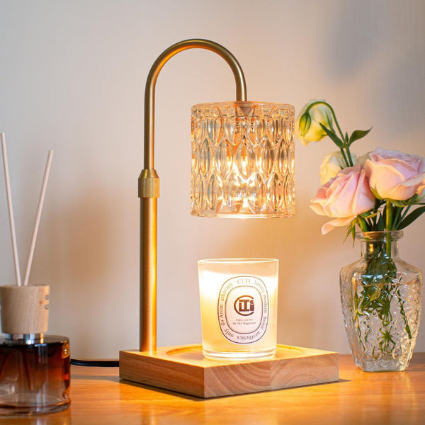 Brushed gold electric candle warmer lamp with textured glass shade over lit candle on wooden base.