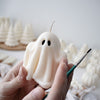 White ghost-shaped candle with black eyes and textured folds, held by two hands with paintbrush nearby.