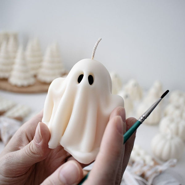 White ghost-shaped candle with black eyes and textured folds, held by two hands with paintbrush nearby.