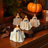 Four small white ghost-shaped candles with black eyes, lit and arranged on a wooden surface with autumn decorations and a.