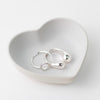 White ceramic heart-shaped tray holding two silver rings and a silver heart pendant.