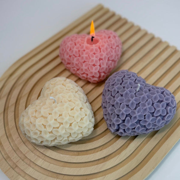 Three heart-shaped candles in pink, purple, and cream with detailed floral texture on a wooden tray.