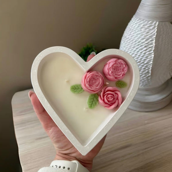 Heart-shaped white ceramic candle with three pink rose decorations and green leaves inside.