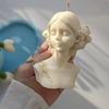 Cream-colored lady sculpture candle with floral hair details and gold accents on chest.