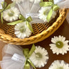 White daisy-shaped candles with green centers wrapped in sheer white fabric and tied with green ribbon in a wicker basket.