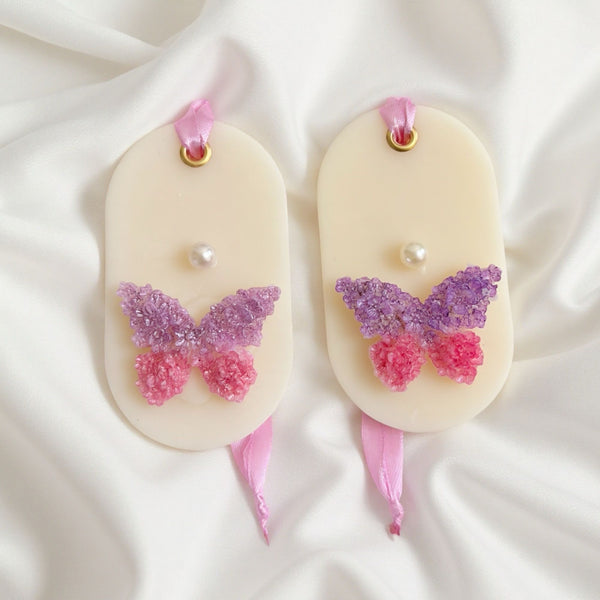 Two cream soy wax hanging sachets with purple and pink glitter butterflies and pearl accents on pink ribbons.