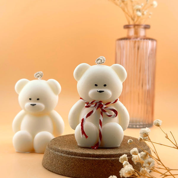 White teddy bear-shaped candle with red and white striped ribbon sitting on cork base against soft orange background.