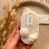 Oval white soy wax sachet with textured seashell designs and small pearls on a wooden tray.