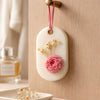 Oval white soy wax sachet with pink flower and dried white blossoms, hanging on wooden knob.